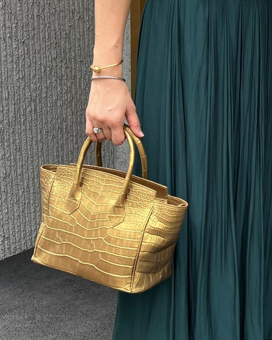 Gold handbag held by a person wearing a teal dress against a gray wall.