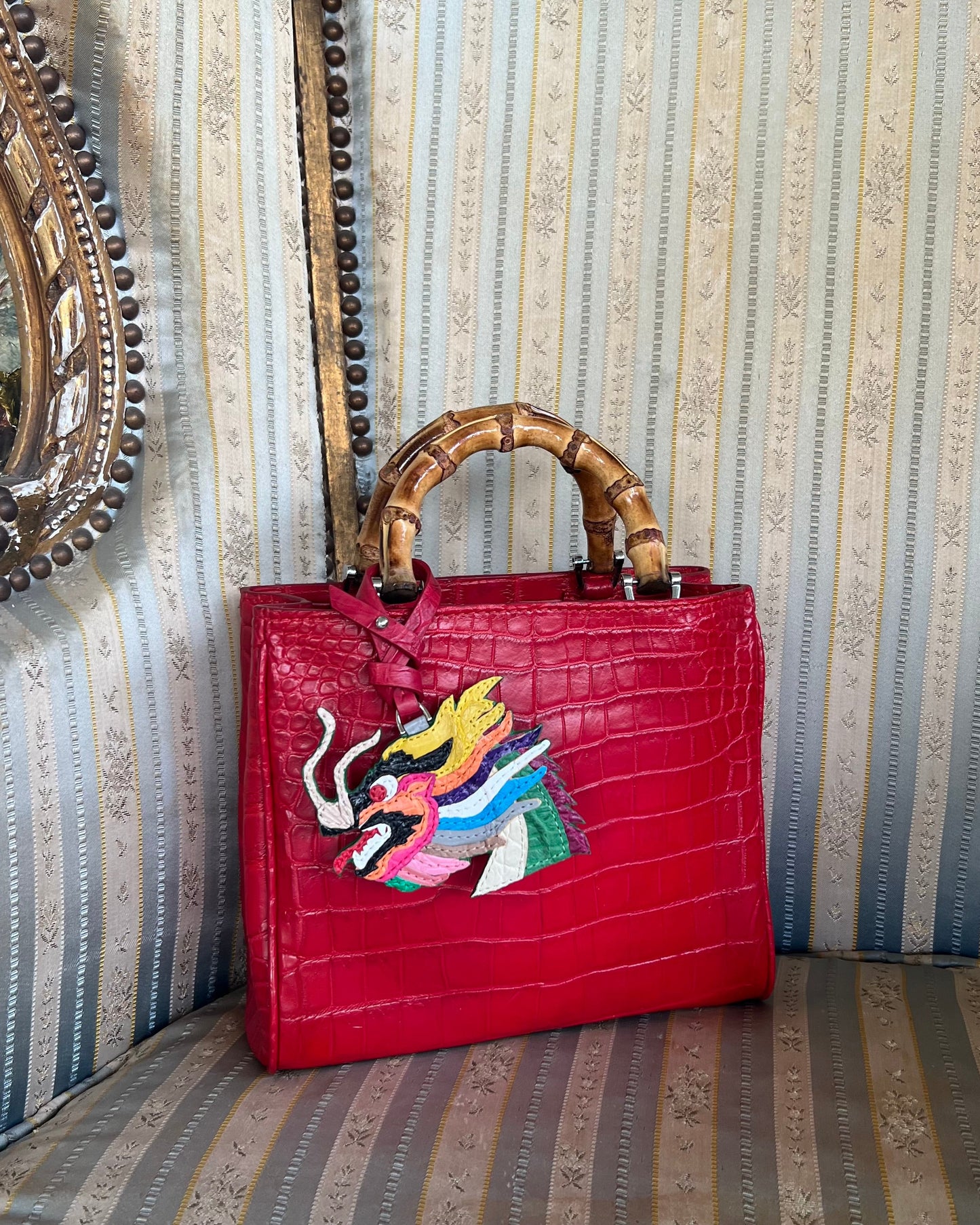 A scarlett red crocodile leather top handle bag with a butterfly embroidery on the front, placed on a striped chair with a decorative background.