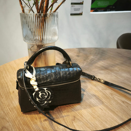 Black handbag with a floral clasp on a wooden table, with a vase of branches in the background.