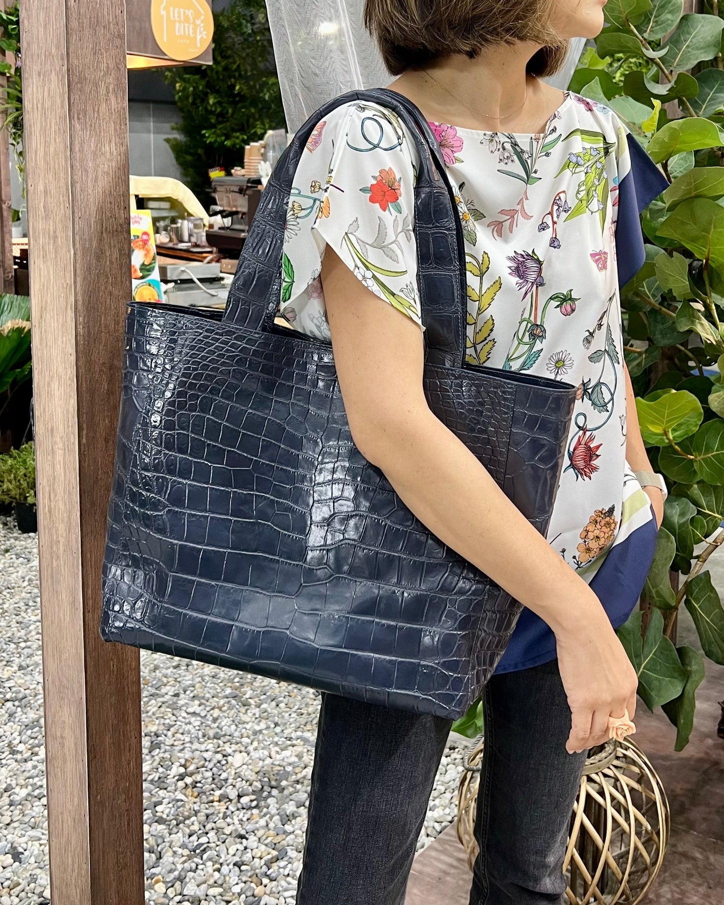 Woman holding a navy blue handbag in an outdoor setting with plants and wooden structures.
