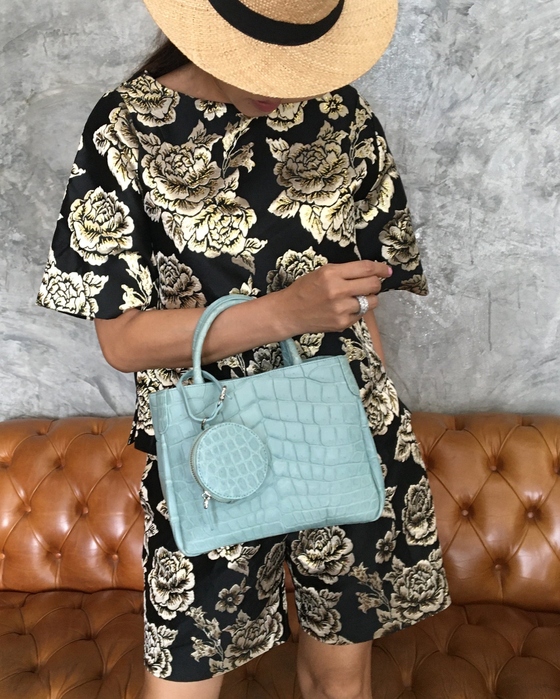 Person standing in front of couch holding a light blue handbag, wearing a black floral dress and straw hat.
