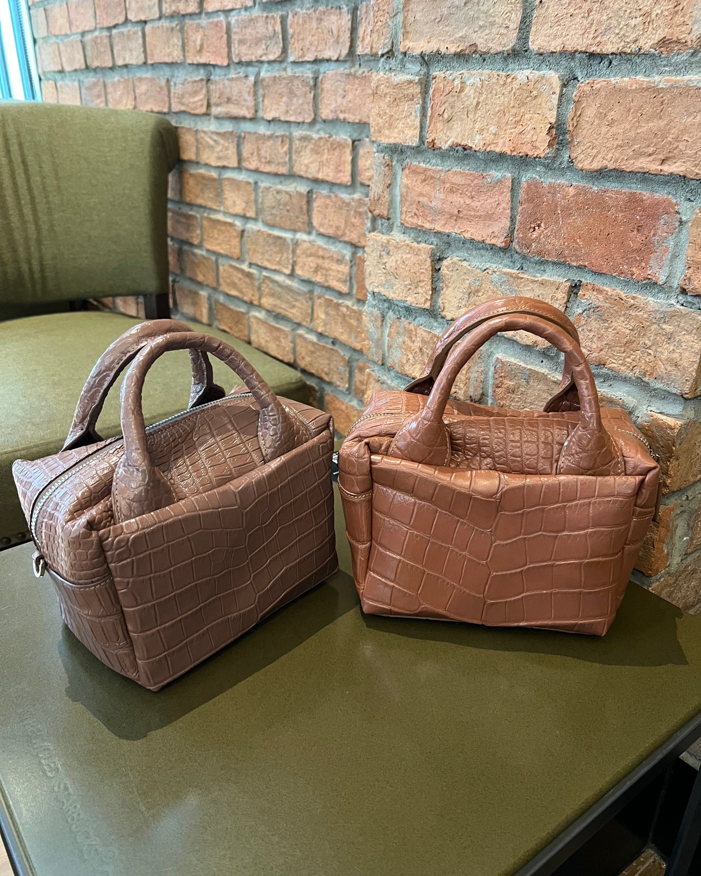 Two brown textured handbags on a green surface with a brick wall background