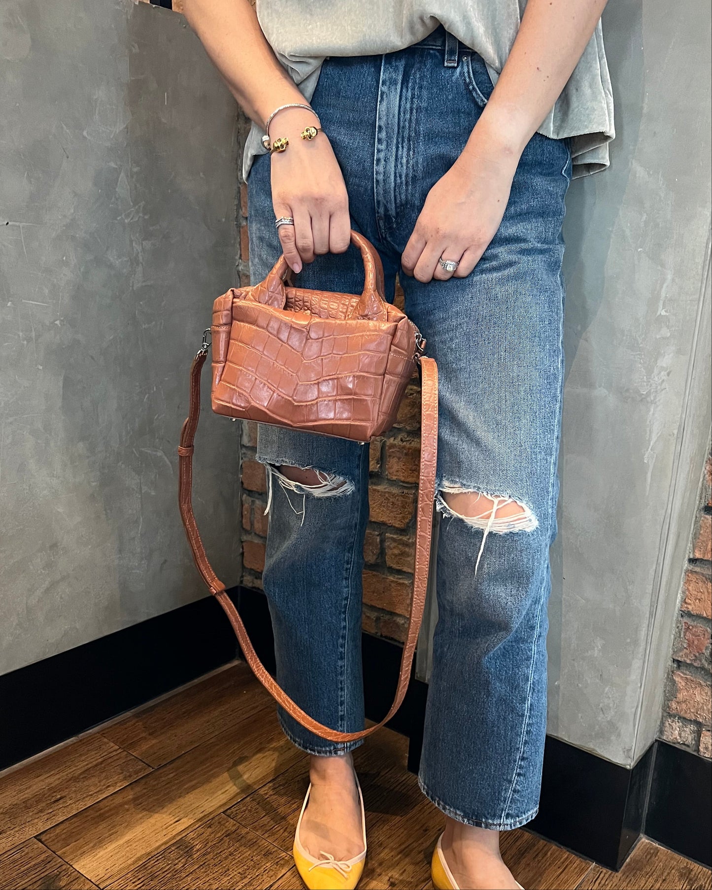 Person sitting on a bench holding a brown leather handbag, wearing ripped jeans and yellow shoes.