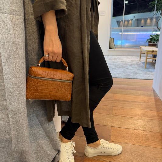 Person sitting on a couch holding a brown leather bag, wearing white sneakers and a long coat.
