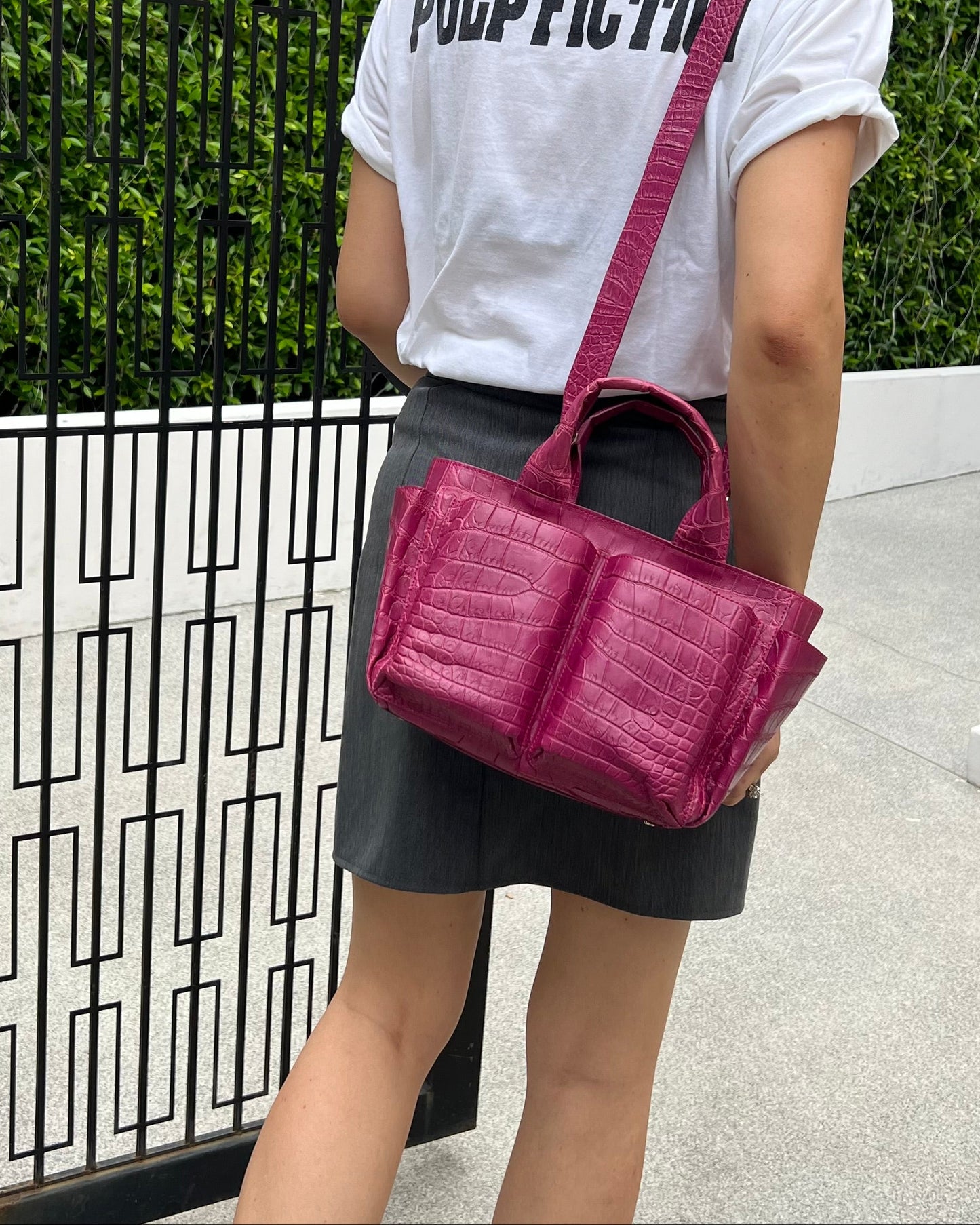 Person holding a pink handbag with 'Pulp Fiction' text on a white shirt, standing near a black metal gate.