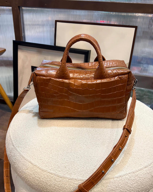 Brown crocodile-patterned handbag on a white cushion with a blurred indoor background