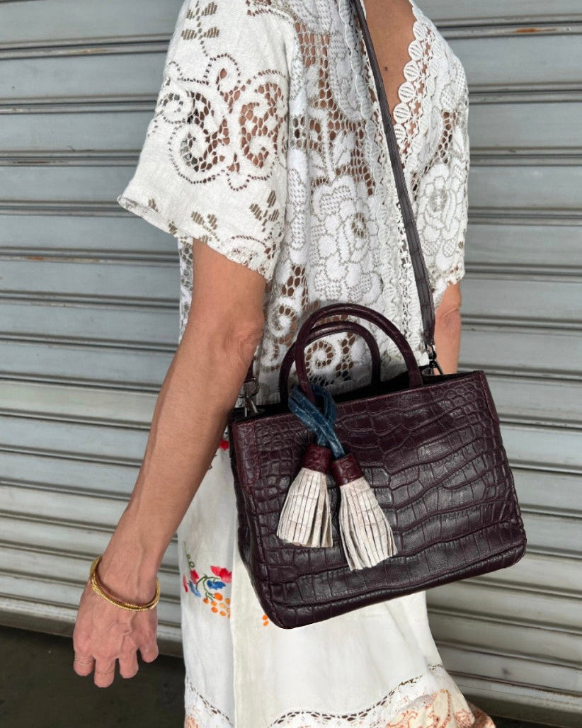 Brown leather handbag with tassels held by a person wearing a white lace top.