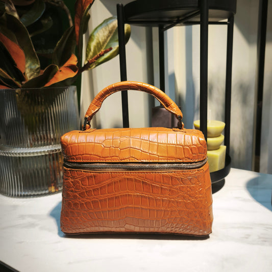 Brown crocodile-patterned handbag on a white surface with decorative items in the background