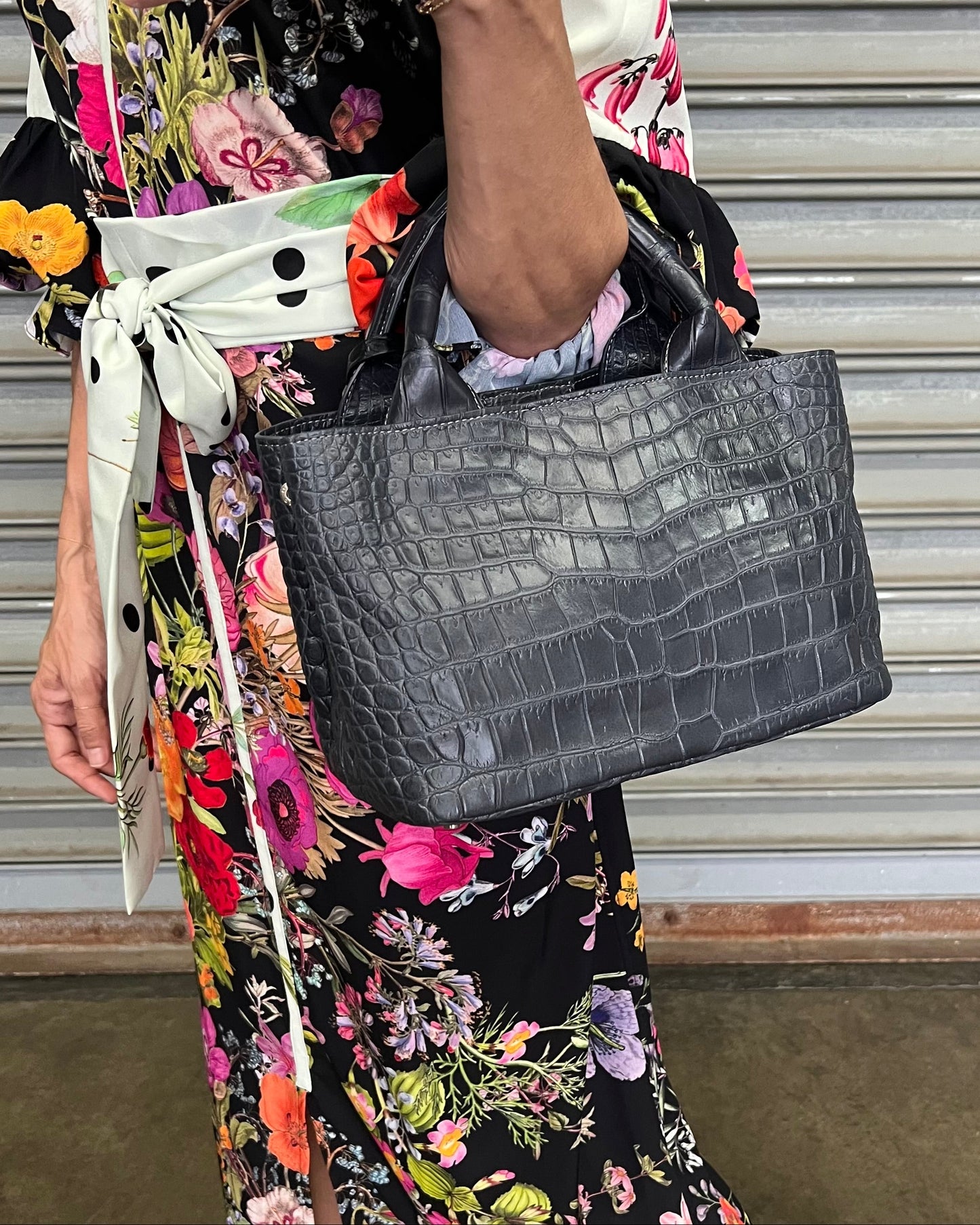 Person holding a black crocodile-patterned handbag with a floral dress in the background