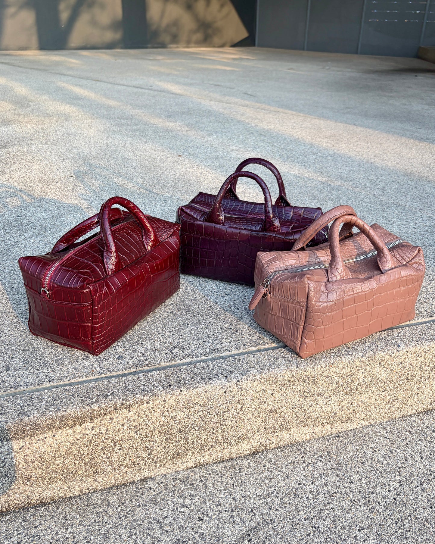 Three leather bags in red, purple, and brown on a concrete surface.