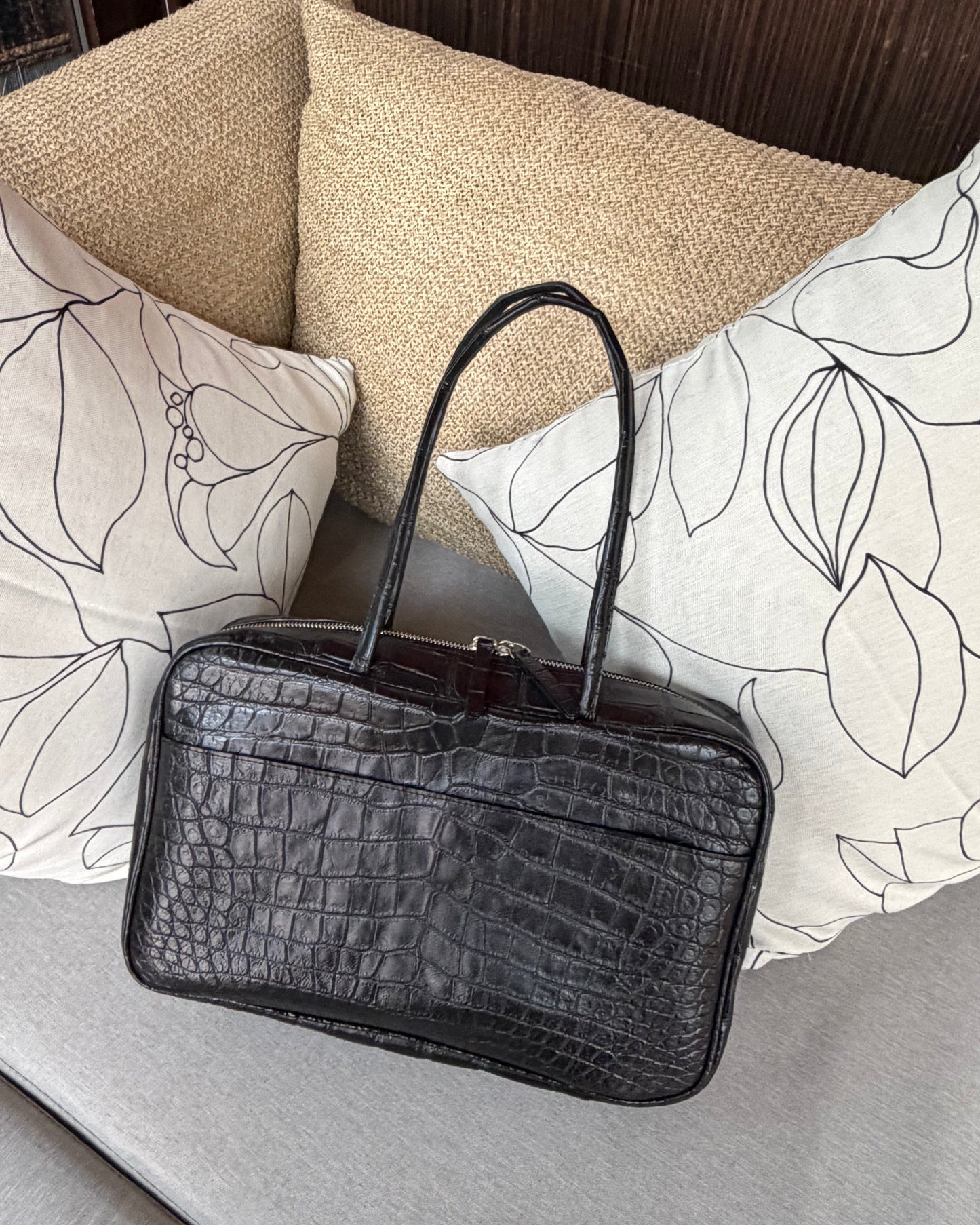 Black crocodile-patterned handbag on a surface with decorative pillows in the background