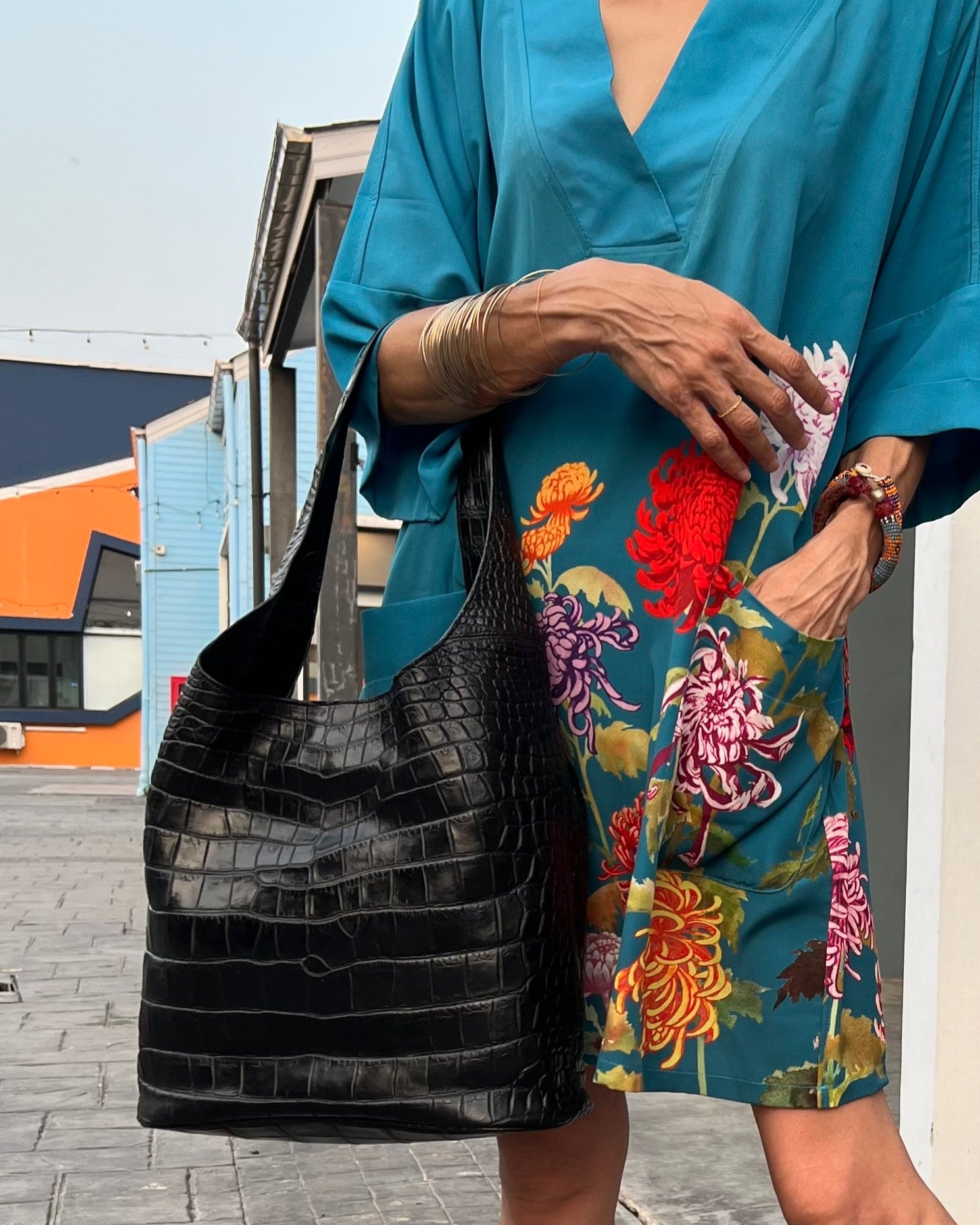 Person wearing a blue top and colorful floral skirt, holding a black bag, standing on a street.