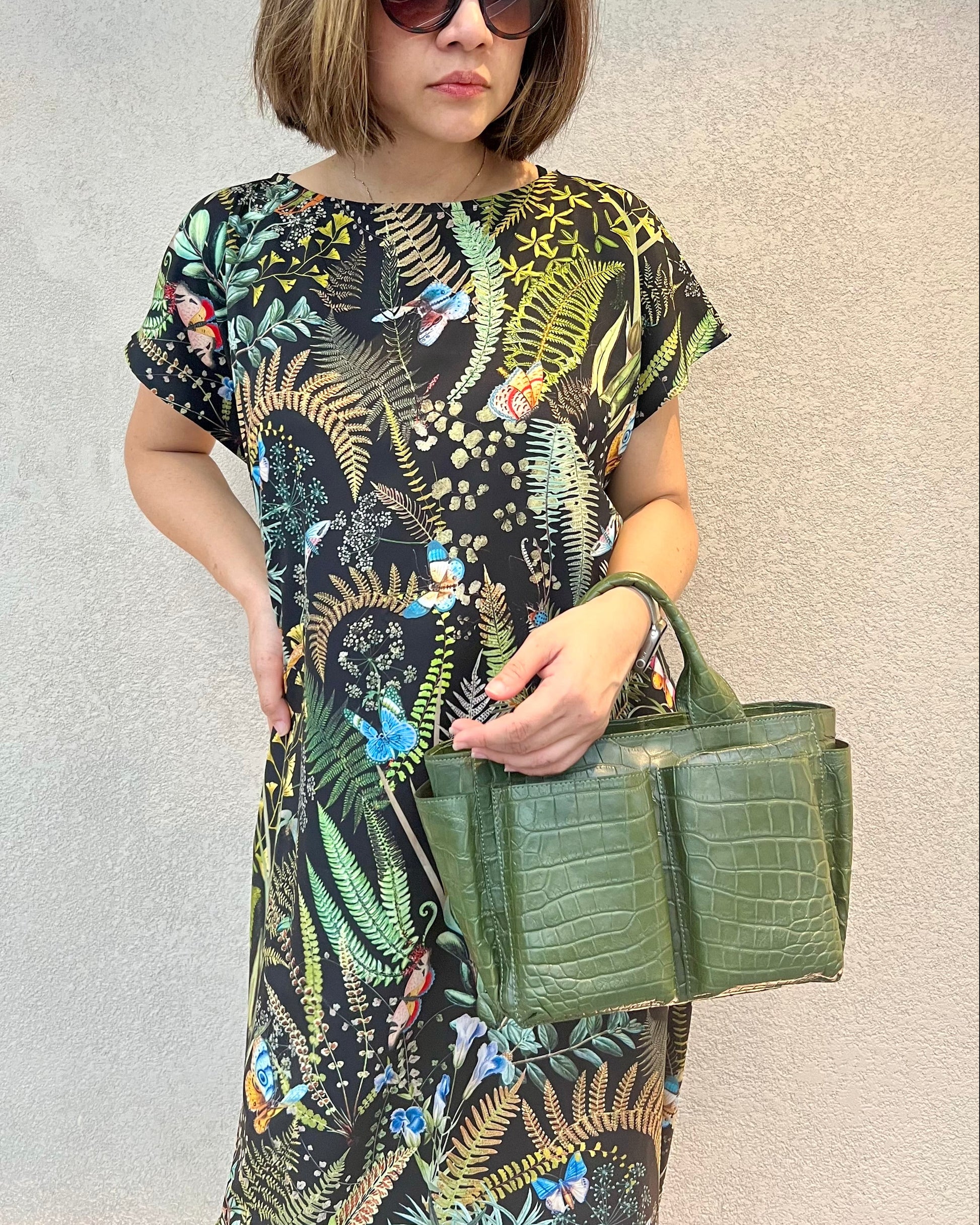 Woman wearing a patterned dress and holding a green handbag against a plain background