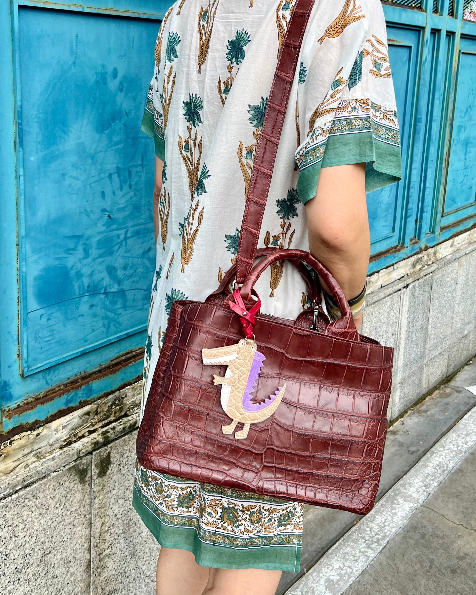 Woman holding a brown handbag with a decorative charm, standing in front of a blue metal fence.