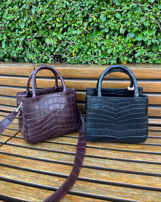 Two handbags, one brown and one black, on a wooden surface with greenery in the background.