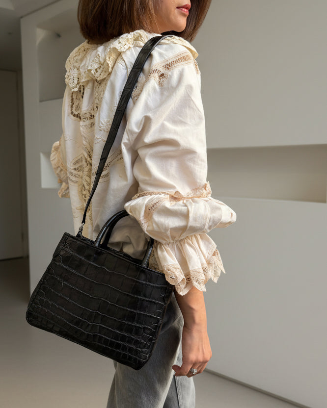 Person wearing a white lace top and gray pants holding a black handbag.