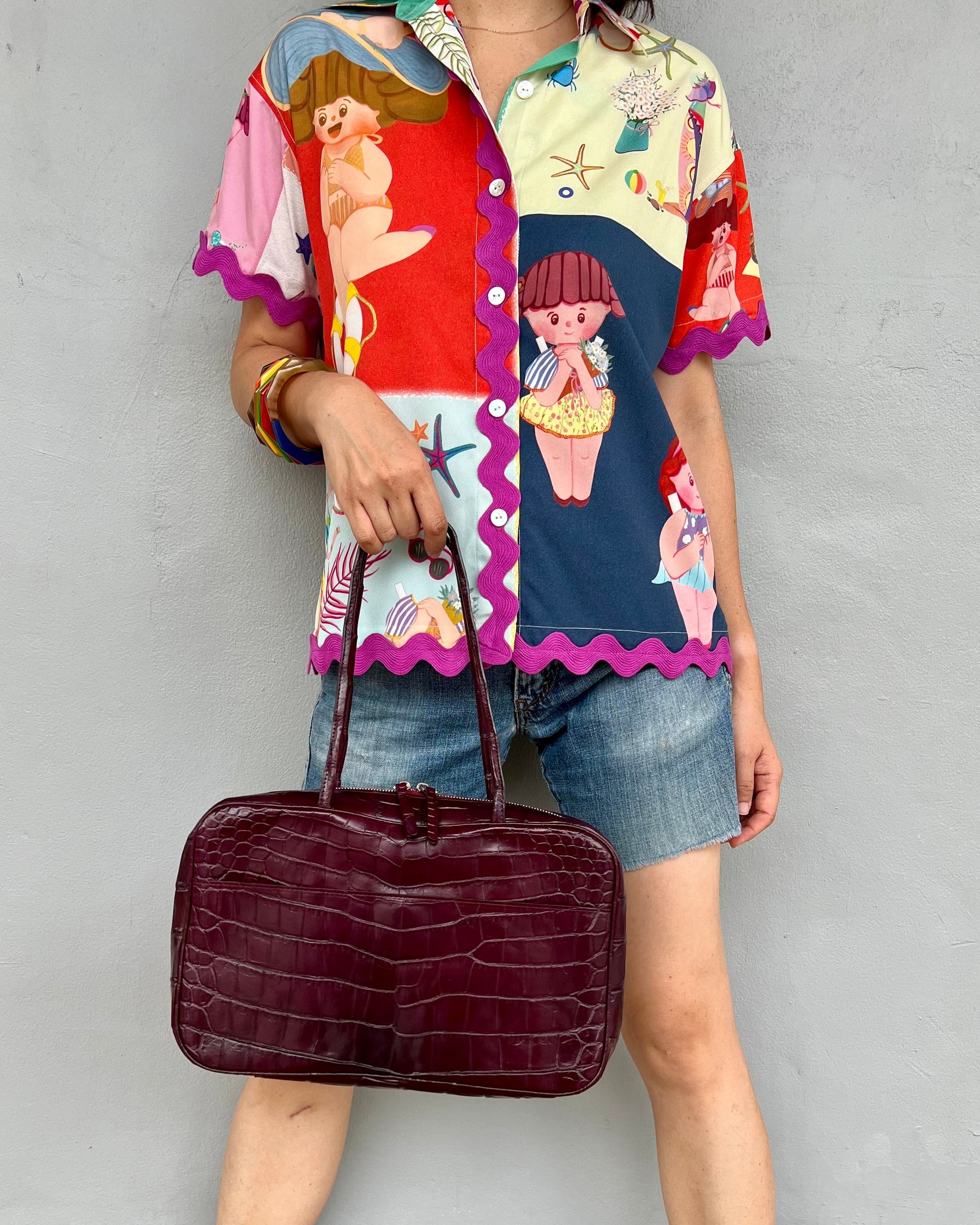 Person wearing a colorful shirt and denim skirt holding a maroon handbag against a plain background