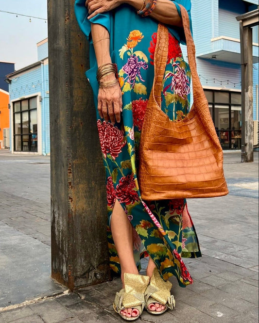 A woman wearing a floral dress and gold open-toe heeled sandals, carrying a brown hobo bag with a crocodile leather texture.