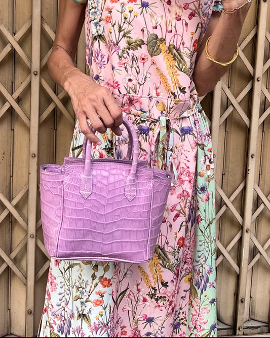 Woman in a floral dress and straw hat holding a purple handbag against a wooden lattice background